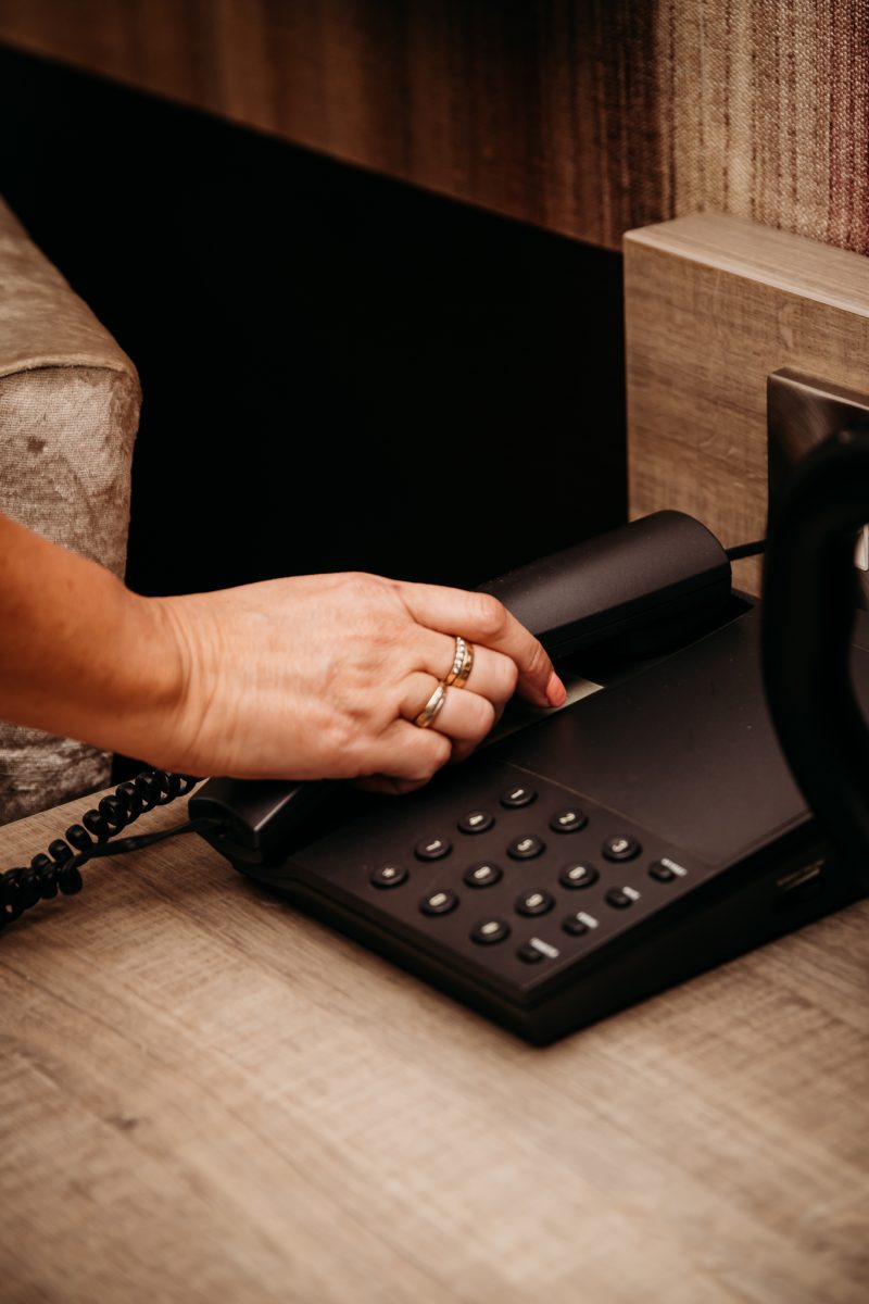 a hand with gold rings about to pick up the receiver of a black phone in a hotel bedroom
