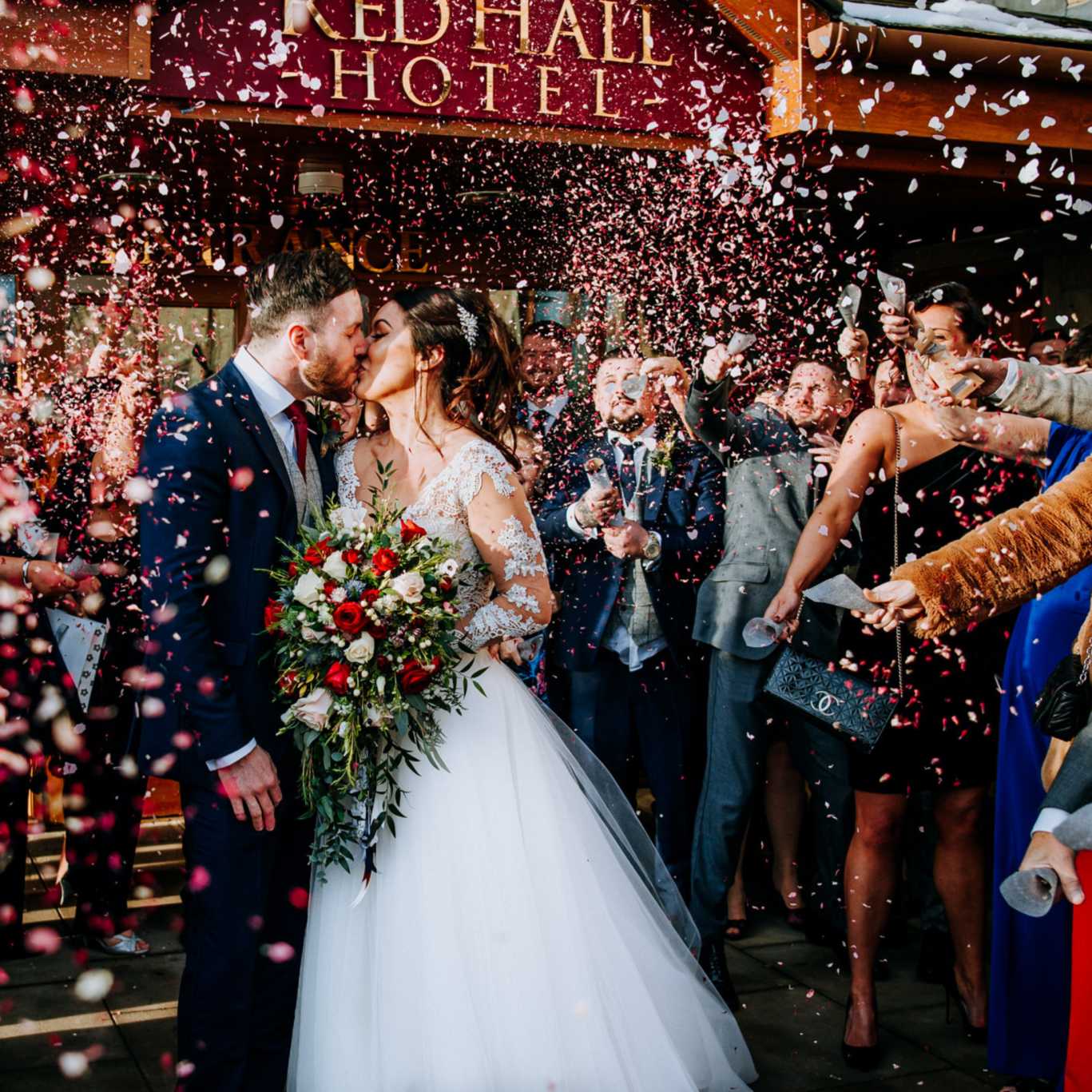 bridge and groom kissing outside red hall hotel with guest throwing red confetti over and around them