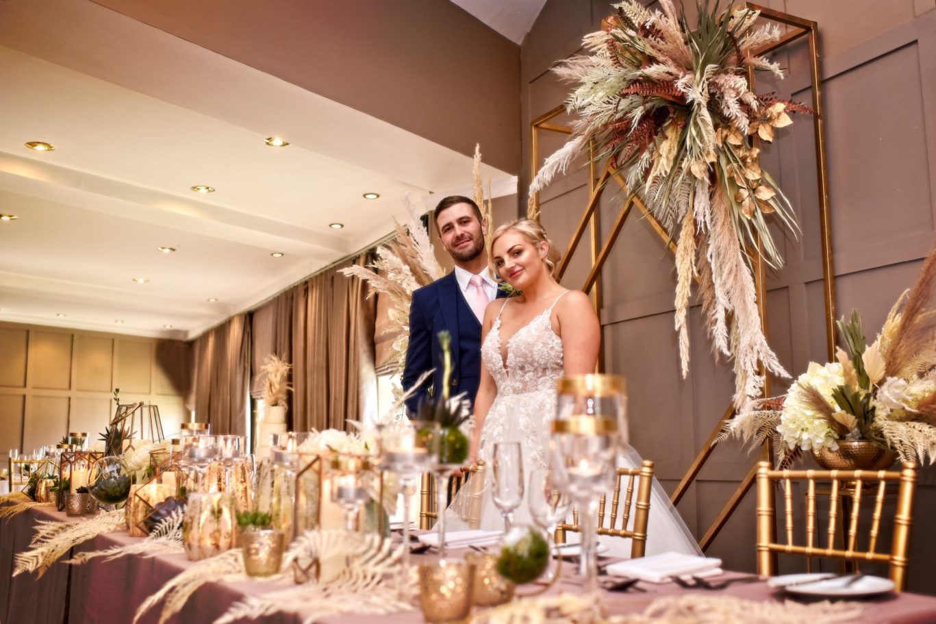 bride and groom standing behind head table decorated with gold and cream pampas grass and candles