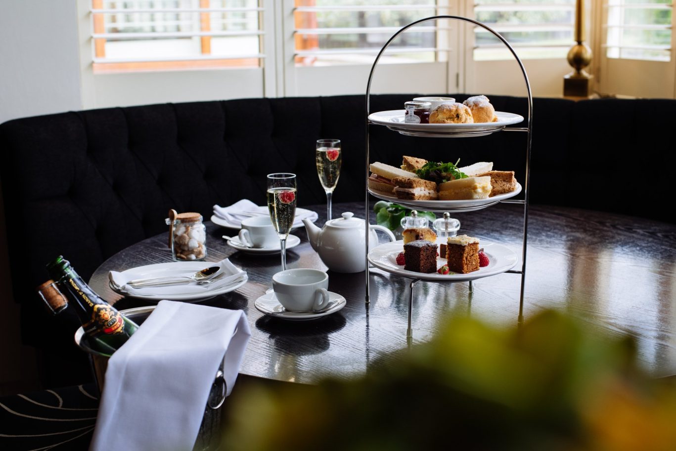 afternoon tea with stand including scones jam and cream on top layer, middle layer has selection of sandwiches and cakes, tea pot and cups, champagne with raspberries on the table