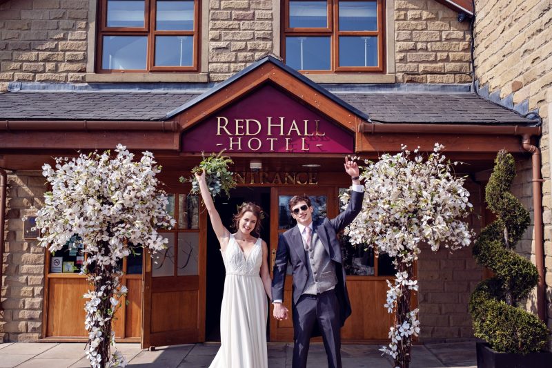 bride and groom standing in front of red hall hotel with arms in the air celebrating