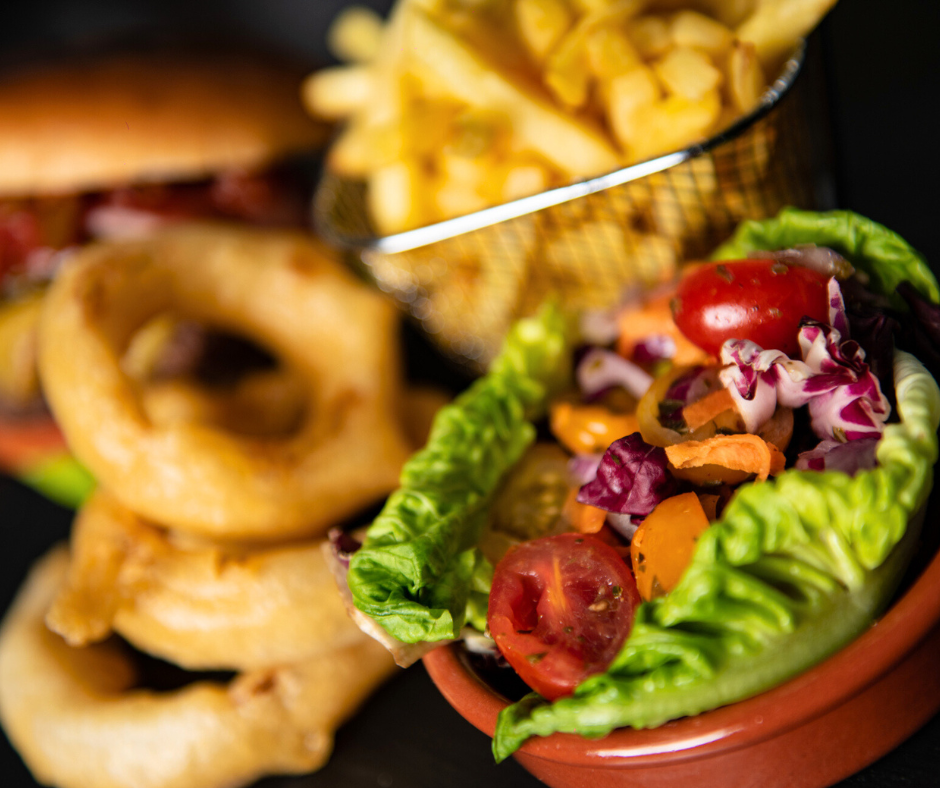 close up of salad, chips and onion rings with a burger in the background