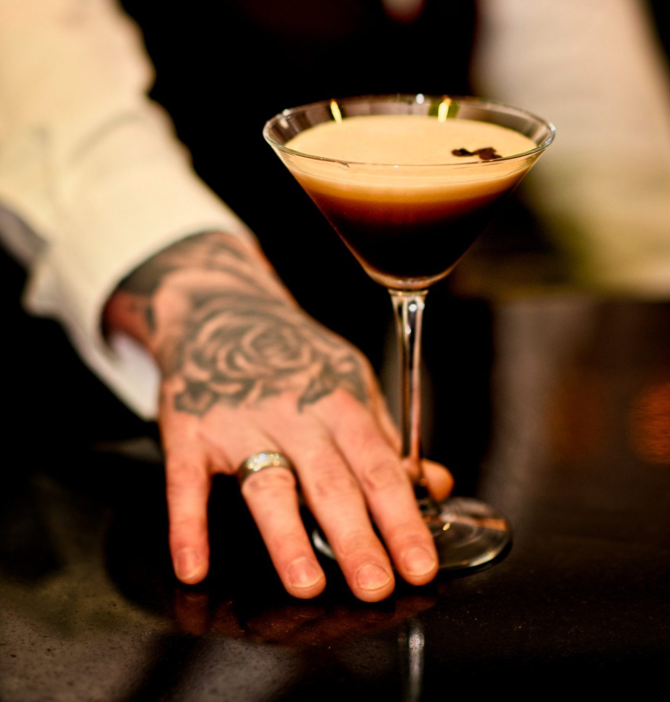 waiters hand pushing an espresso martini toward camera, waiter has a rose tattoo on hand and wedding ring