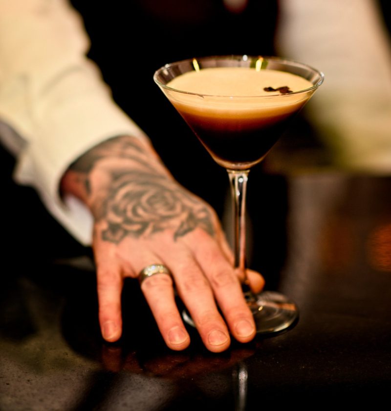 waiters hand pushing an espresso martini toward camera, waiter has a rose tattoo on hand and wedding ring