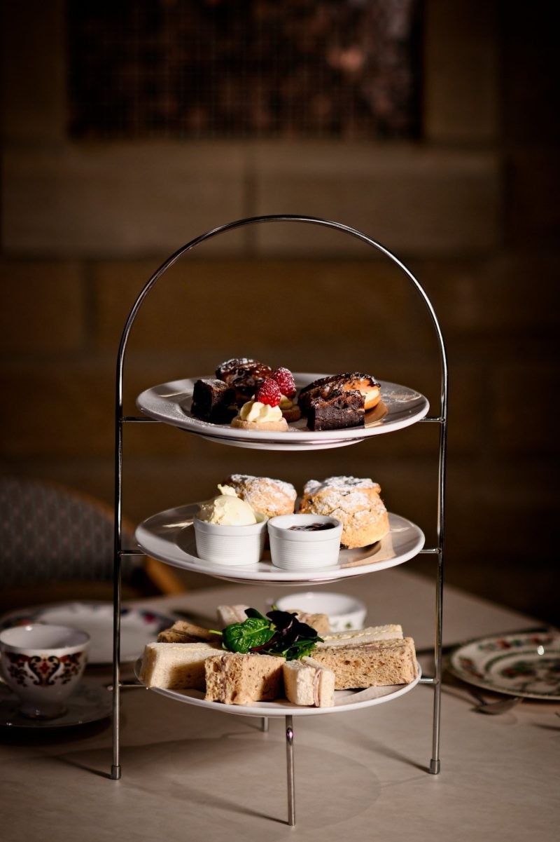 afternoon tea stand with cakes and brownies on the top later, scones with cream and jam in the middle, and the bottom layer with an assortment of sandwiches on white and brown bread