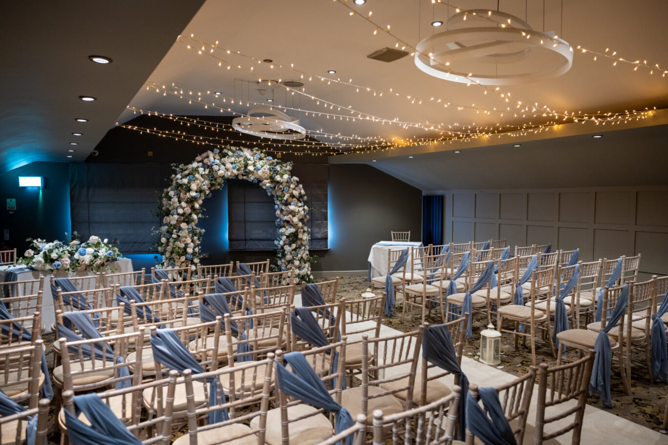 Modern and beautifully furnished wedding ceremony room with statement flower arch, sashed chairs for around fifty wedding guests, whimsical fairy lights, and a white carpeted aisle at Red Hall Hotel near Bury.