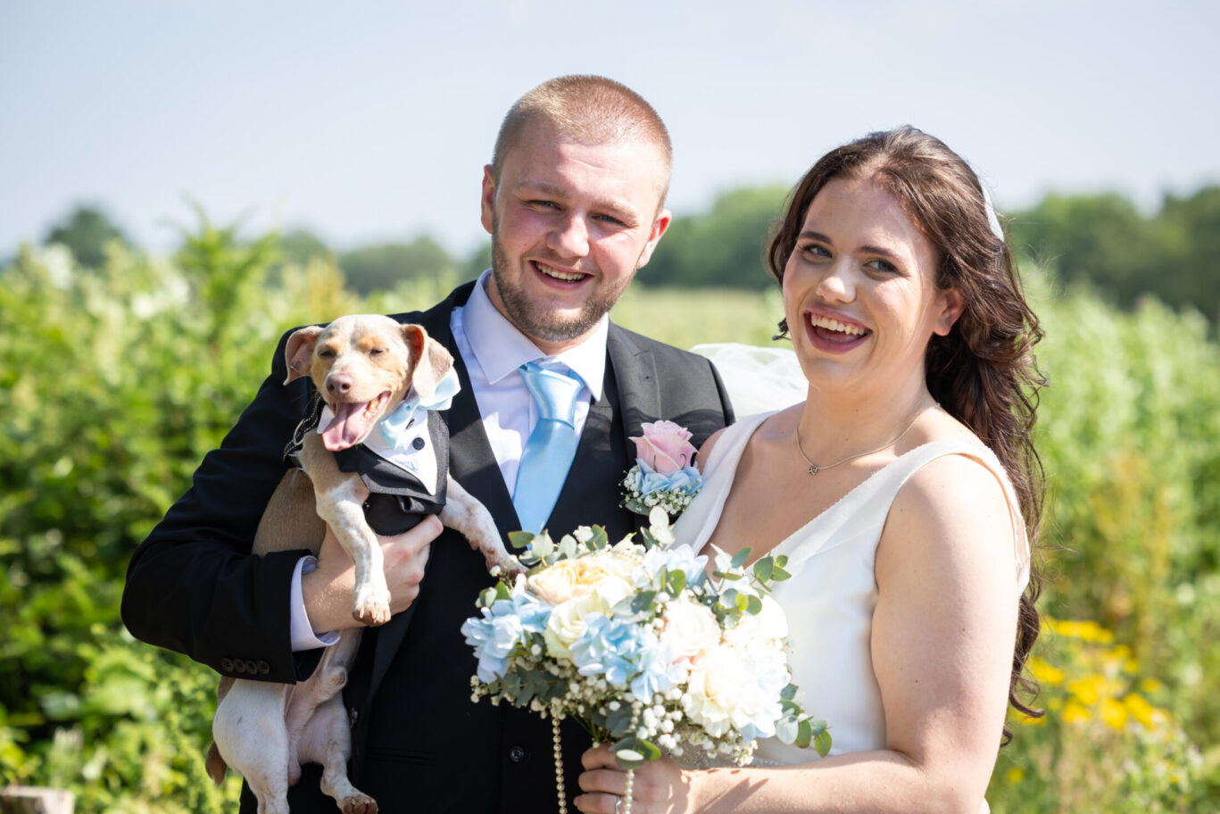 Bride and groom smiling and holding a small dog in a tuxedo and bow tie as part of Red Hall Hotel's outdoor dog-friendly wedding space in Bury.