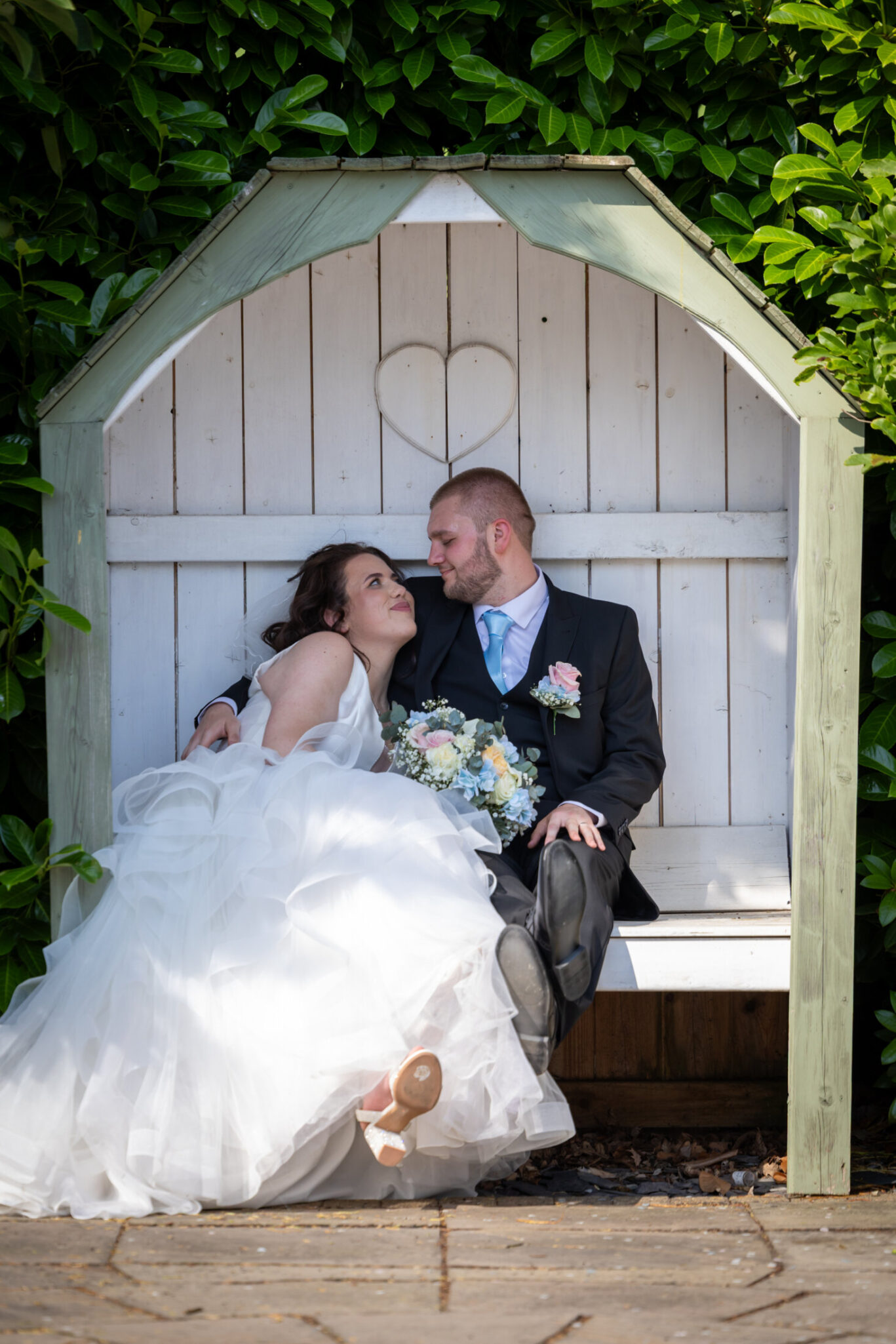 Bride and groom smiling and embracing underneath a shaded bench area with a white heart, in part of Red Hall Hotel's outdoor wedding venue space, Bury.