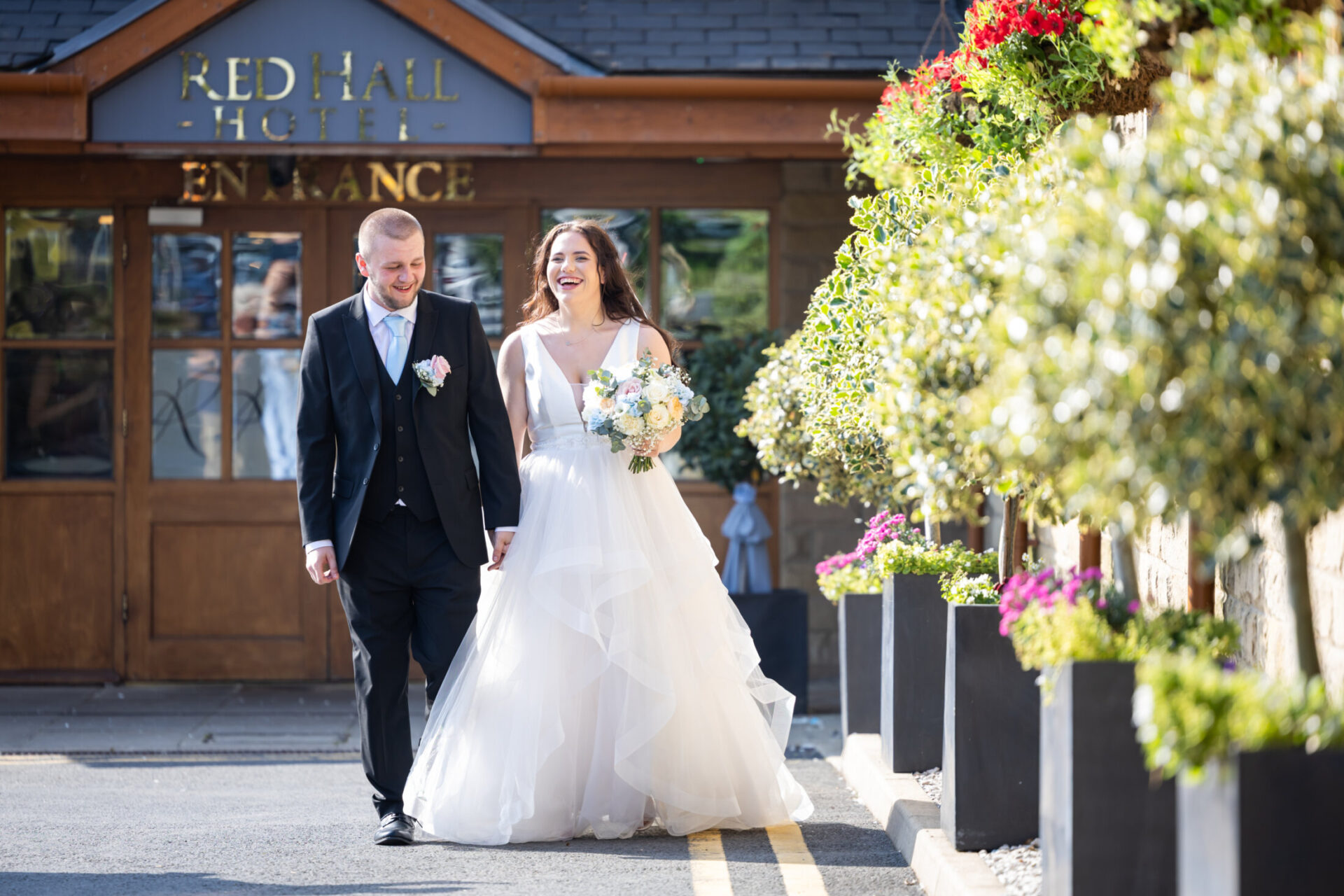 Bride and groom walking hand in hand smiling in front of the foliage and entrance of the Red Hall Hotel in Ramsbottom.