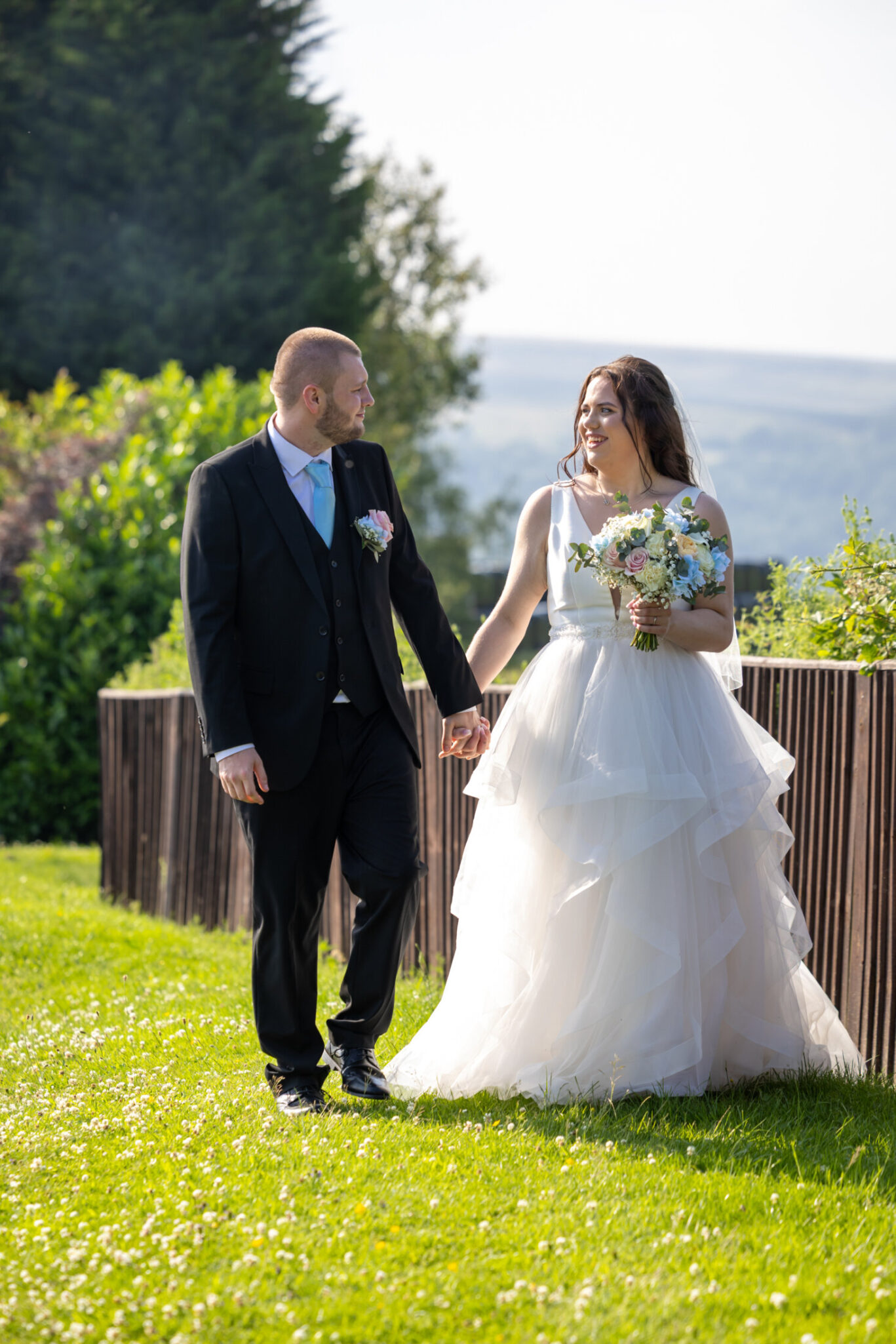 Bride and groom walking hand in hand across the grassy outdoor wedding space at the Red Hall Hotel in Bury.