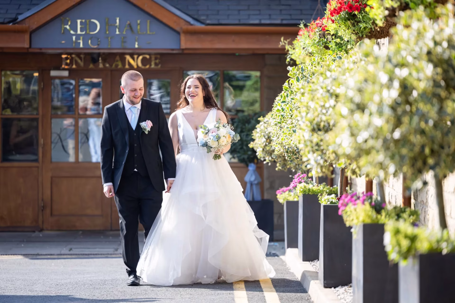 Bride and groom walking hand in hand smiling in front of the foliage and entrance of the Red Hall Hotel in Ramsbottom.